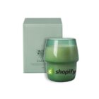 A green candle in a glass holder is placed next to a matching green box. The glass has the word shopify with a small green shopping bag logo on it.