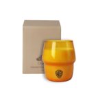 Orange glass candle holder with a black logo, placed beside a brown cardboard box featuring a small graphic. The box has a handwritten note style font with a sketch. Candle holder and box are on a plain white background.