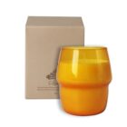 A yellow glass candle holder with a textured middle section, placed in front of a brown cardboard box displaying an illustration and text.