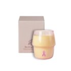 A pink candle with a pink ribbon symbol on the side sits in front of a white box. The candle is in a round glass container and symbolizes breast cancer awareness.