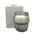 A white candle in a gray glass holder placed in front of a white box. The box features a red logo with text, and a bowl illustration with small text underneath.