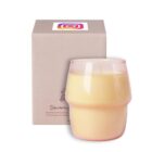 A beige box with a colorful logo on top stands next to a lit, cream-colored candle in a round glass holder. The candles wax is smooth and fills most of the holder.