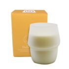 A white, frosted glass candle is placed next to a yellow box. The box features minimal design elements and text. The candle has a modern, layered design and appears unused.