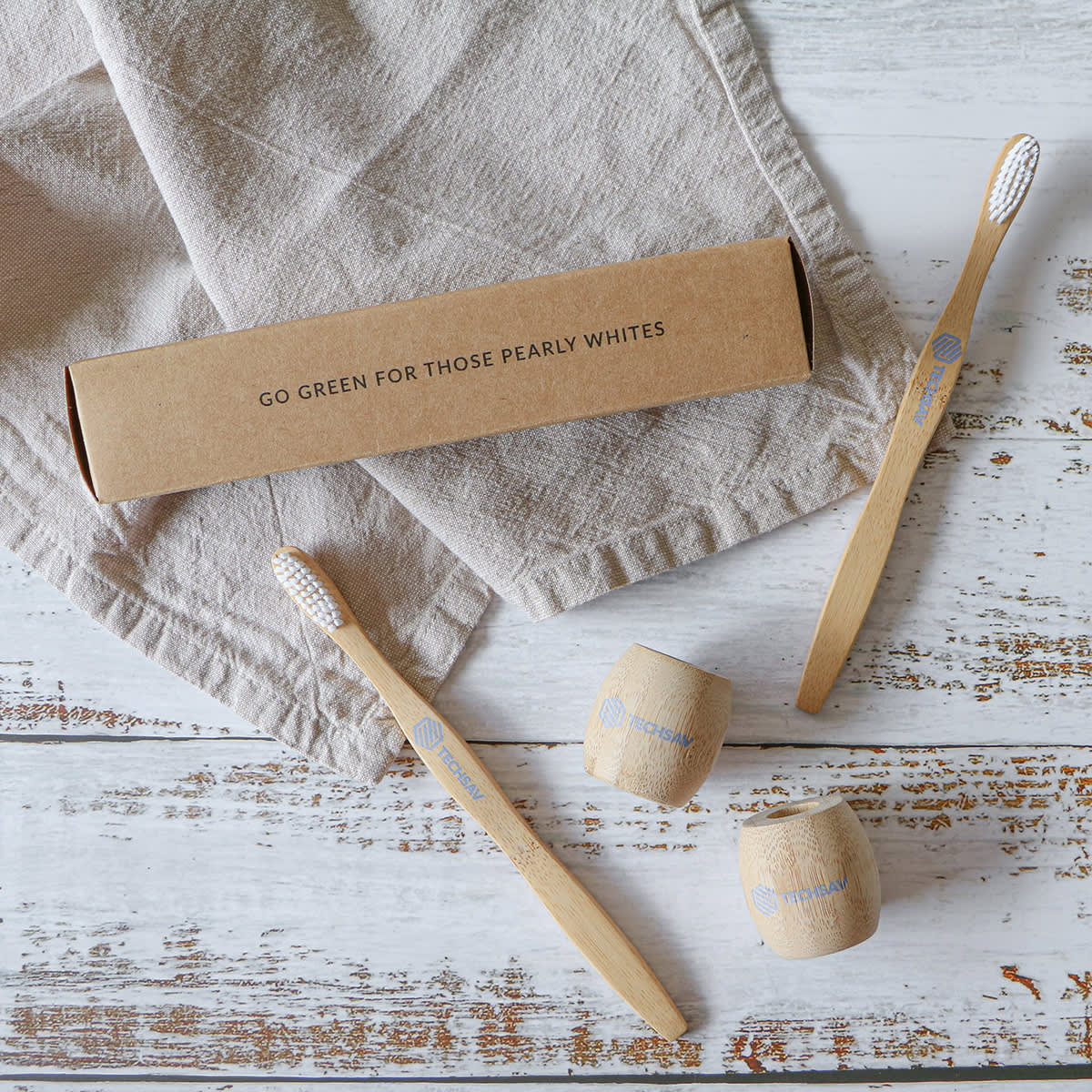 Three bamboo toothbrushes with white bristles are on a rustic wooden surface next to two wooden holders and a cardboard box. The box has a message: Go green for those pearly whites. A beige cloth is partially visible underneath.