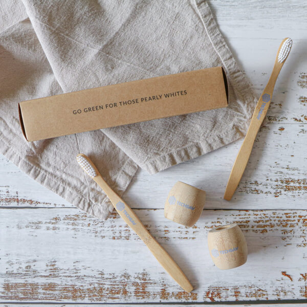 Two bamboo toothbrushes with white bristles and accompanying wooden holders are placed on a rustic white wooden surface. A brown cardboard box with the text “Go Green For Those Pearly Whites” is nearby, resting on a beige cloth.