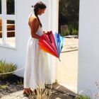 A woman in a long white dress stands outside a building, holding a colorful umbrella with red, orange, yellow, blue, and purple sections. She is looking down, with a braided hairstyle, and there is a chair in the background.