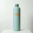 A tall, cylindrical water bottle with a matte light green finish stands on a white surface against a plain background. The bottle features the SoundCloud logo in orange on its front.