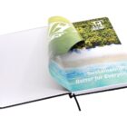 Open notebook with a cover featuring a nature-themed image of a beach and forest. The text on the cover reads Sustainability Better for Everyone and includes a logo at the top. A black ribbon bookmark is visible.
