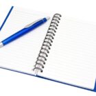 An open spiral-bound notebook with lined pages on a white background. A blue pen is placed diagonally across the pages. The notebook has a blue cover with a black clasp.