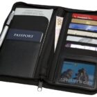 Open travel wallet with a passport, pen, various credit and membership cards, currency notes, and a card labeled travellers case. The wallet is black with a zipper.