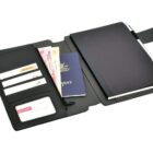 A black leather travel organizer is open to reveal multiple compartments. Inside are a passport, a notebook, several business cards, and what appears to be a boarding pass. A pen is clipped to the notebook.