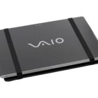 A closed black Sony Vaio laptop with a silver logo on the lid. The laptop is secured with two black elastic straps. The edges are sleek, and the overall design is minimalist and modern.