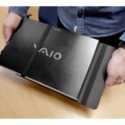 A person holds a black Sony Vaio laptop enclosed in a protective case. The person is wearing a blue shirt, and the laptop is partially opened with visible branding. The background includes a wooden table surface.