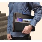 A person in a blue button-up shirt holds a leather portfolio. The portfolio has pockets containing a smartphone, pen, and a notebook. They are standing next to a staircase.