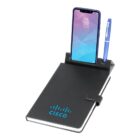 A black notebook with a Cisco logo, featuring a blue pen and a smartphone holder integrated into its cover. A smartphone is placed in the holder, displaying a geometric lock screen.