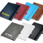 Seven colorful notebooks are displayed in two rows. The top row has black, red, turquoise, and brown notebooks. The bottom row features a gray notebook with a logo, and plain white and black notebooks. Each has a ribbon bookmark and elastic closure.