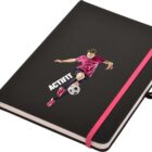 A black notebook with a pink elastic band features an illustration of a soccer player kicking a ball. The word ACTIFIT is printed on the cover above the ball.