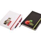 Two notebooks are shown. The first is white with a cat image and the word PAWS in red, featuring a red pen. The second is black with a red apple image and the text THE HEALTH GROUP in green, accompanied by a green elastic band.