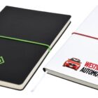 Two notebooks with elastic bands. The black notebook has a green band and the AUSGLASS logo. The white notebook has a red band, a car graphic, and the WESTSIDE AUTOMOTIVE logo. A pen is attached to the white notebook.