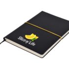 A black notebook with Slice of Life printed in white text on the cover, alongside an illustration of sliced citrus fruit. The notebook is secured with a thin yellow elastic band and has a ribbon bookmark.