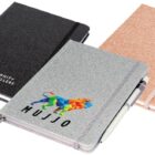 Three notebooks with sleek covers and elastic bands. The black notebook has a geometric logo, the grey one features a colorful lion and Mujjo, and the pink one is plain. A pen is attached to the pink and grey notebooks.