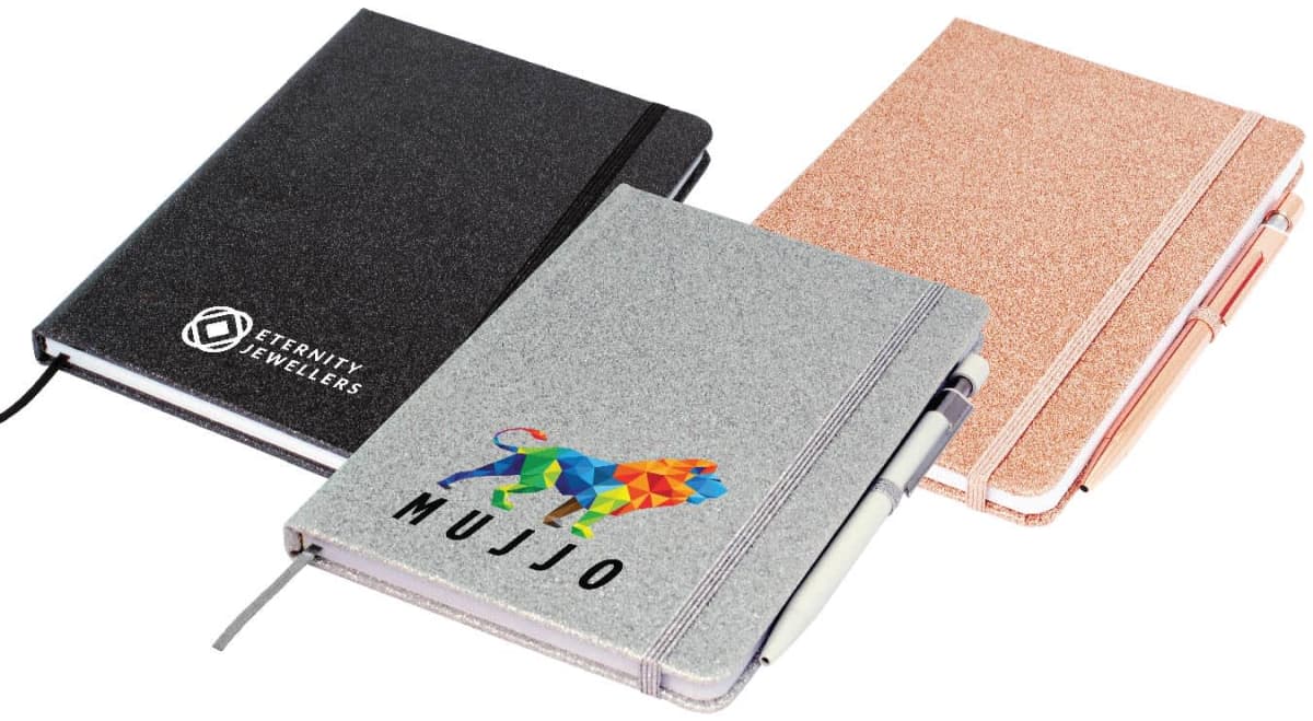 Three notebooks with sleek covers and elastic bands. The black notebook has a geometric logo, the grey one features a colorful lion and Mujjo, and the pink one is plain. A pen is attached to the pink and grey notebooks.