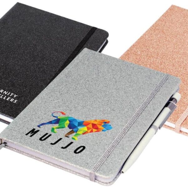 Three closed notebooks are displayed in a row: a black one with Eternity Jewellers in white, a silver one with a colorful lion design and MUJJO, and a rose gold one. Each has an elastic band, and the silver one holds a pen.