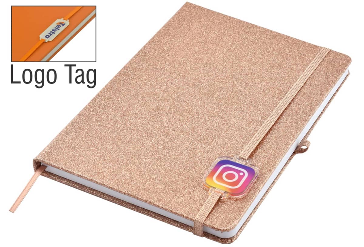 A glittery gold notebook with a beige elastic band featuring the Instagram logo. A small tagged logo is attached to the band. The notebook includes a matching ribbon bookmark.