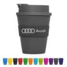 Gray reusable cup with Audi logo and Cup 2 Go text, featuring a silicone grip. The cup is placed above a row of ten colorful cups in various shades, including yellow, purple, red, green, and blue.
