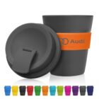 A group of colorful reusable cups with lids. One black cup with an orange band and logo reading Audi stands prominently in the foreground. Other cups in various colors like yellow, red, blue, pink, and green are lined up at the bottom.