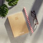 A tan textured notebook lies on a stone surface beside tortoiseshell glasses, a red and white striped straw, and a green monstera leaf. Sunlight casts shadows across the scene.