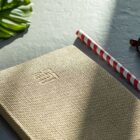 A textured beige notebook with a subtle embossed design on the cover is placed on a gray surface. Next to it is a red and white striped pencil and a small part of a green leaf.
