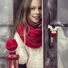 Girl in a white sweater and red scarf smiles while holding a red lantern. She stands by a snow-dusted door with a reindeer ornament on the handle, creating a cozy, festive atmosphere.