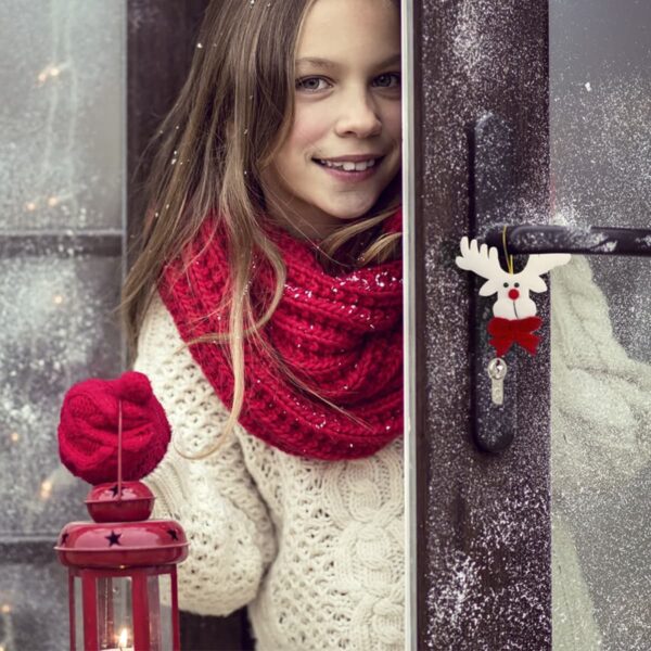 Girl with long hair wearing a red scarf and gloves stands by a frosty door. She holds a red lantern and smiles. The door has a reindeer-shaped decoration on the handle. Snowflakes are falling.