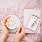 A person with manicured nails holds a cup of marshmallow-topped hot chocolate. Nearby, on a pink plush surface, is a pink notebook with a small clipboard. A vial labeled Hueligold lies on top.