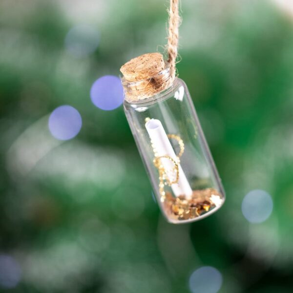 A small glass bottle with a cork stopper hanging by a twine against a blurred green background with bokeh lights. Inside the bottle is a rolled-up scroll and shiny gold confetti.