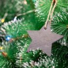 A black star-shaped ornament hangs on a Christmas tree branch with vibrant green needles and a dusting of white snow. Multicolored lights softly illuminate the background, adding a festive touch.