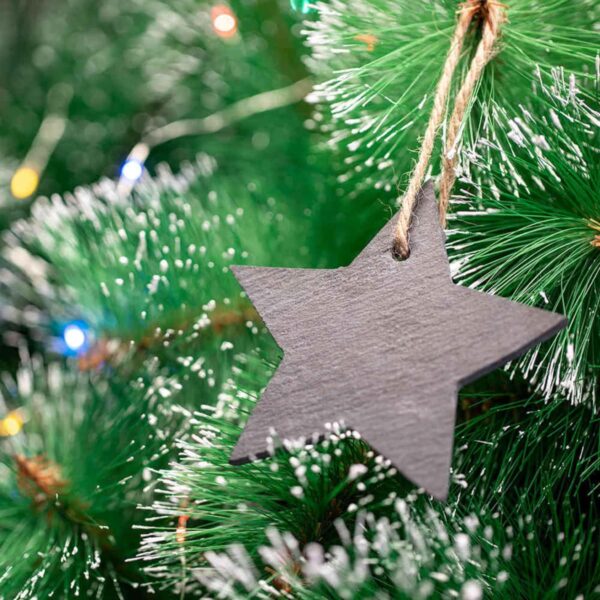 A black wooden star ornament hangs from a jute string on a lush green Christmas tree with snow-like frosted tips. Multicolored lights softly glow in the background, enhancing the festive atmosphere.