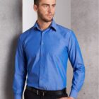 Man wearing a blue button-up shirt and black trousers stands against a textured gray wall. He has short brown hair and a beard, looking to his right with a neutral expression.