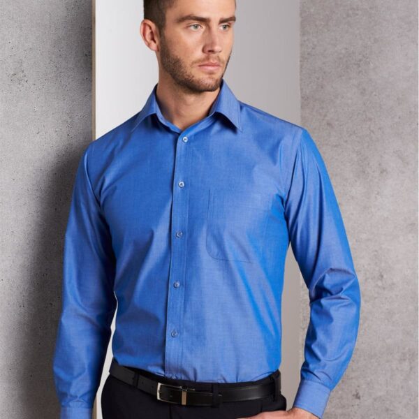 Man wearing a blue button-up shirt and black pants standing against a gray wall, looking to the side.