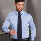Man in a light blue dress shirt with a navy tie and black pants leans casually with one hand on a table. He wears a watch and looks downwards, standing against a gray background.
