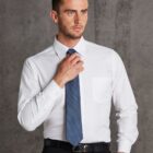 Man wearing a white dress shirt and blue tie, adjusting his tie with one hand. He has short hair and a trimmed beard. He is standing against a textured gray background, looking slightly to the side.