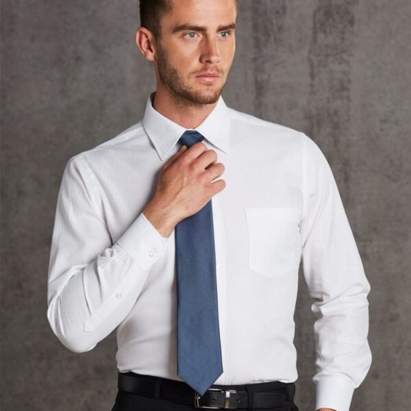 Man wearing a white dress shirt and blue tie, adjusting his tie with one hand. He has short hair and a trimmed beard. He is standing against a textured gray background, looking slightly to the side.
