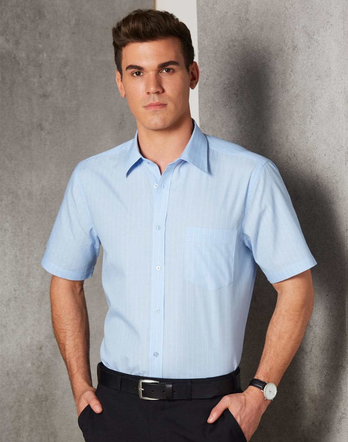 A person is standing against a gray textured wall, dressed in a light blue short-sleeved shirt and dark pants, with hands in pockets. The person is wearing a black belt and a wristwatch, looking directly at the camera.