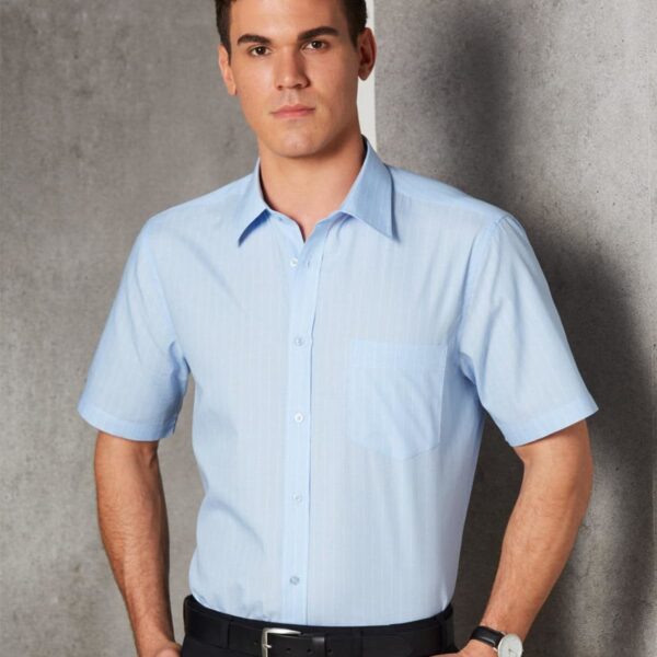 A person wearing a light blue short-sleeve button-up shirt stands with hands in pockets. They have short dark hair and are wearing a watch on the left wrist. The background is a textured gray wall.