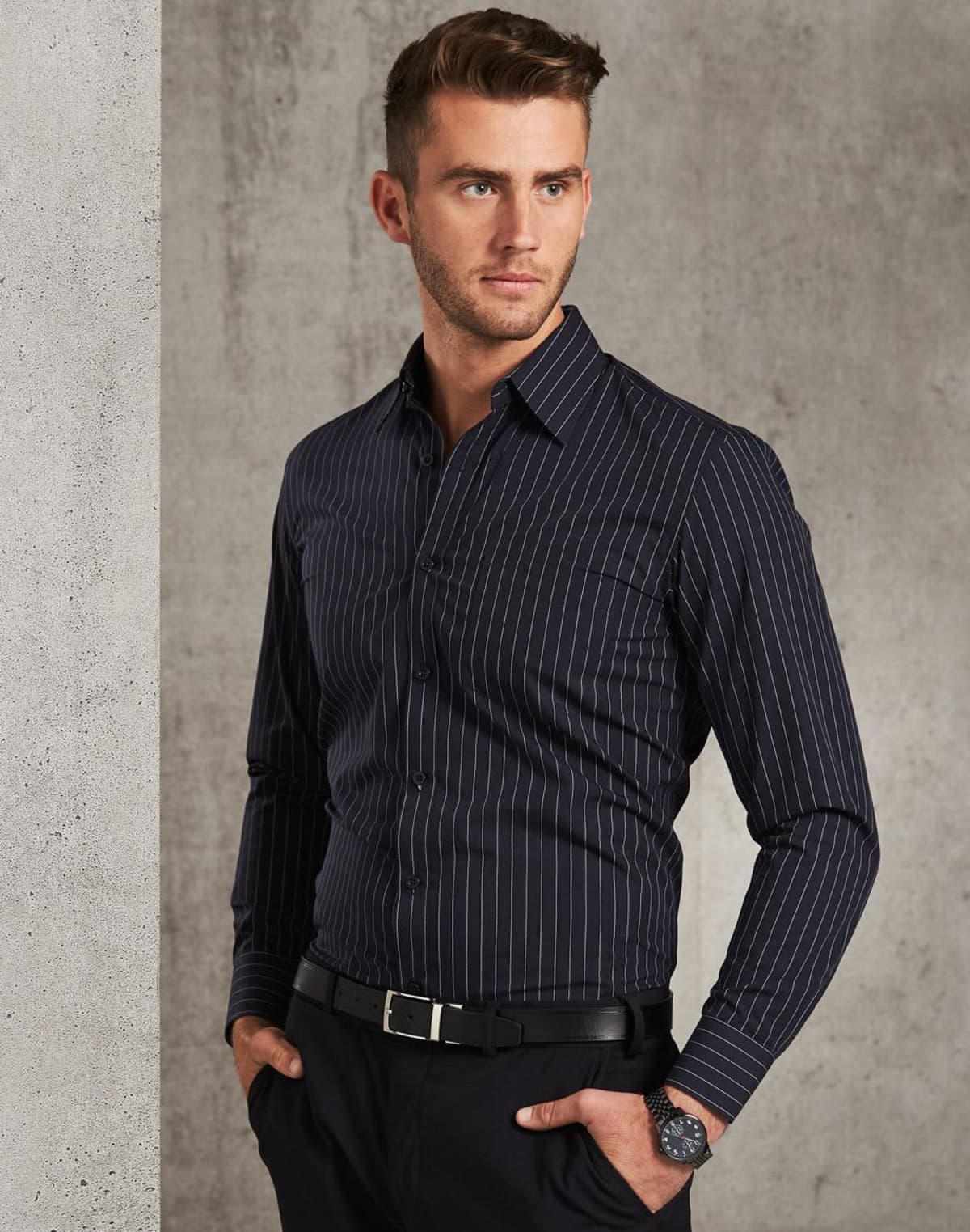 A man in a fitted, dark pinstripe shirt stands with his hands in his pockets, looking to the side. He wears a black belt and a watch, and is set against a textured gray wall.