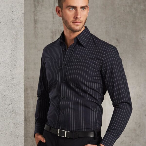 A man in a tailored, dark pinstripe shirt and black pants stands against a textured gray wall. He wears a black belt and a wristwatch, looking thoughtfully into the distance. His hair is styled neatly.