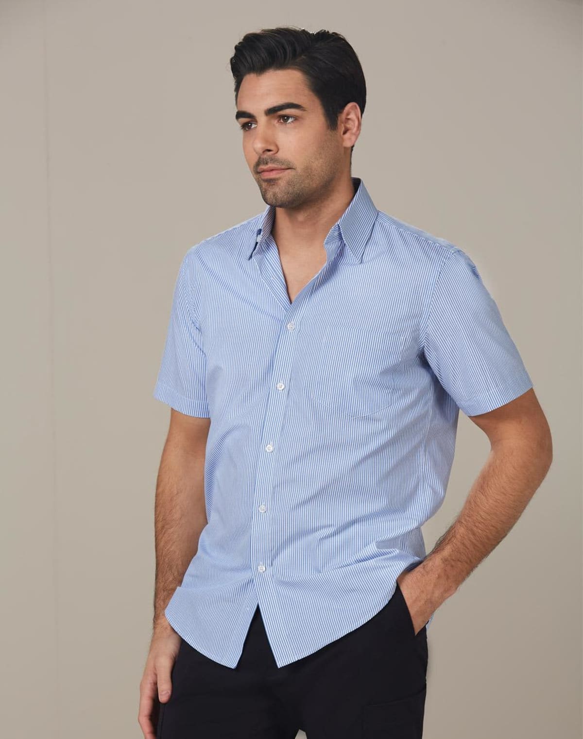 Man wearing a light blue, short-sleeved button-down shirt with subtle stripes, standing with hands in pockets against a neutral background.