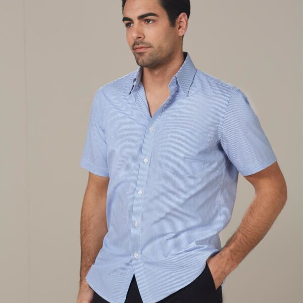 A person wearing a light blue, short-sleeved button-up shirt stands against a beige background. They have short dark hair, and their left hand is in their black pants pocket. The person is looking slightly to the side.