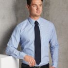 A man in a blue striped shirt, black tie, and black pants stands leaning against a light-colored wall. He has short brown hair and looks to the side with a neutral expression. The background is a textured gray wall.